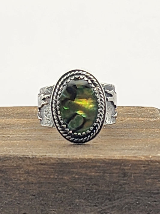 A sterling silver ring with a textured band and gator accents, oval abalone shell with twisted wire border, displayed on a wooden block against a white background.