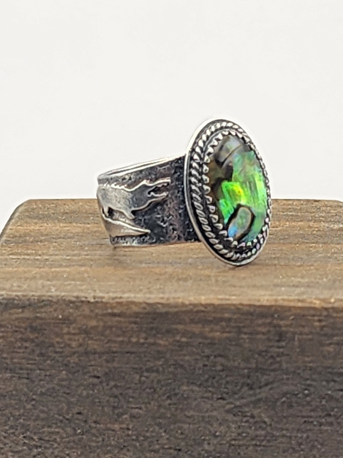 A side view of a sterling silver ring with a textured band and gator accents, oval abalone shell with twisted wire border, displayed on a wooden block against a white background.