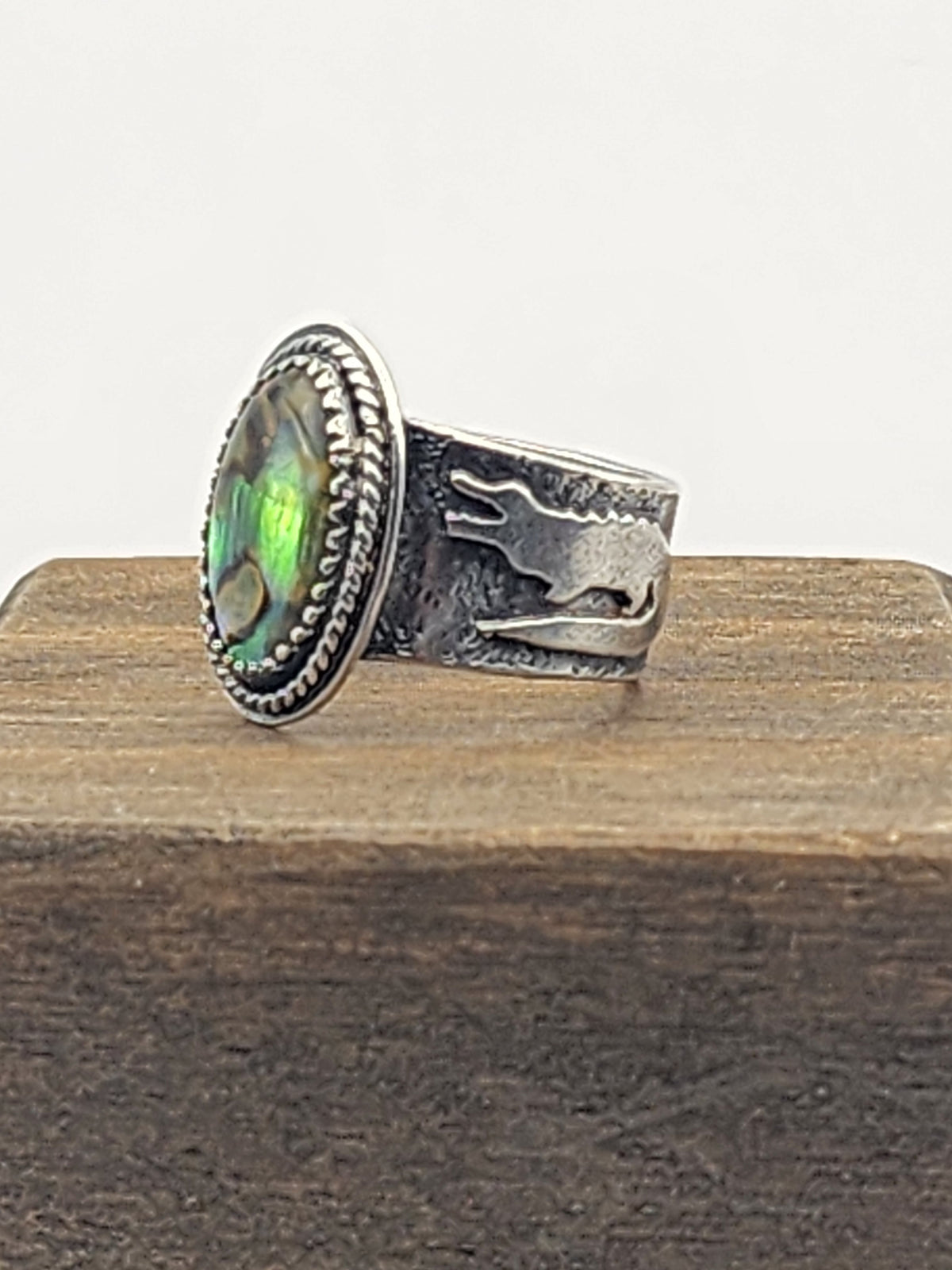 A side view of a sterling silver ring with a textured band and gator accents, oval abalone shell with twisted wire border, displayed on a wooden block against a white background.