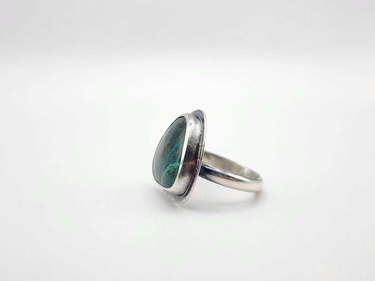 A sterling silver ring featuring a large, irregularly triangle shaped green stone with dark inclusions, set in a polished bezel on a white background.
