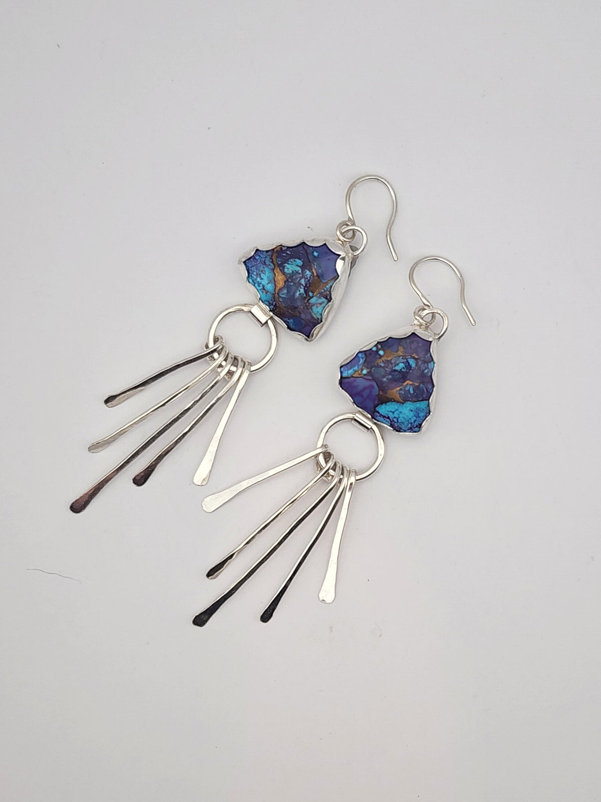 A pair of sterling silver dangle earrings featuring triangular shaped blue and purple mohave turquoise stone pendants with sterling silver fringe, displayed on a neutral light gray background.