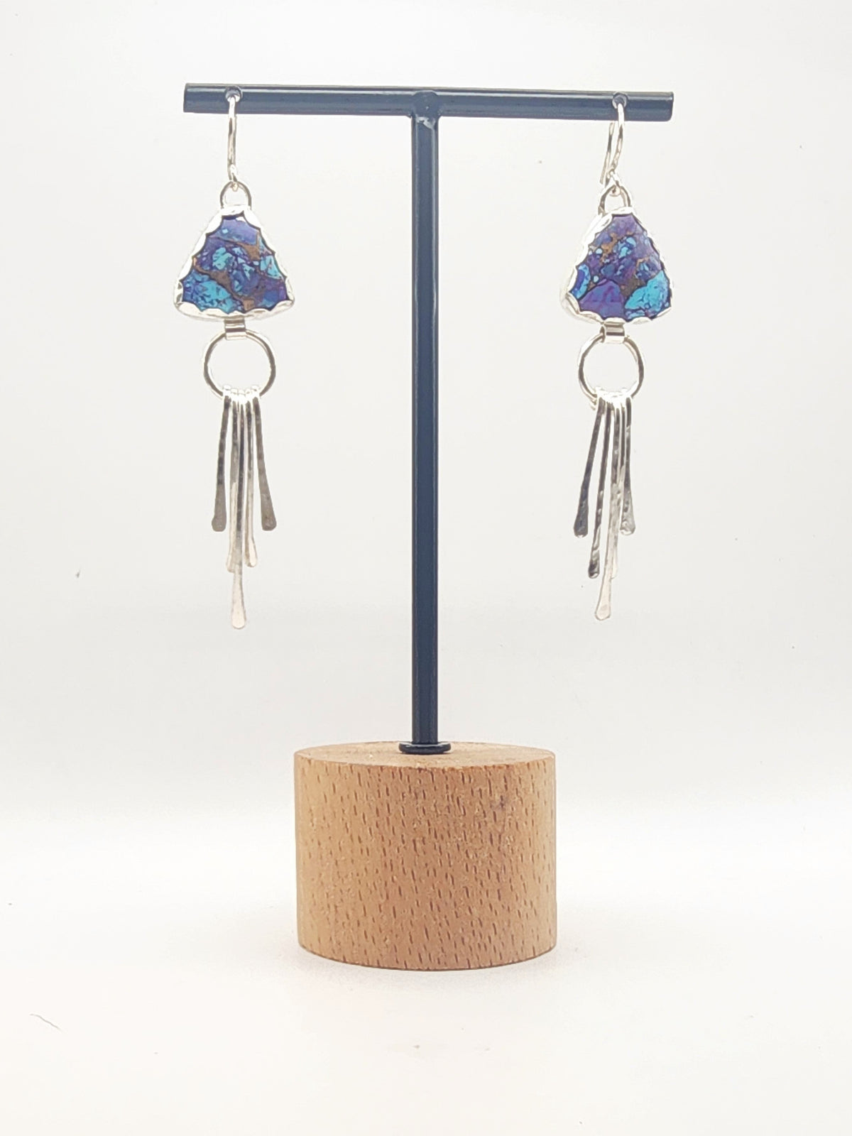 A pair of dangling earrings displayed on a black T-stand with a round wooden base against a plain white background. Each earring has a triangular shaped purple/blue mohave matrix stone, a silver ring, and four dangling silver fringe strips.