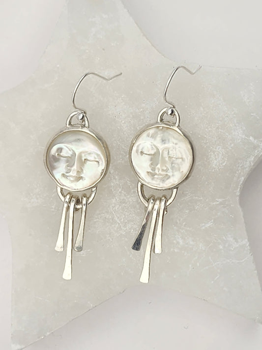 Pair of sterling silver earrings with round moon face stones from mother of pearl with three sterling silver hammered fringes, sitting on white stone display.
