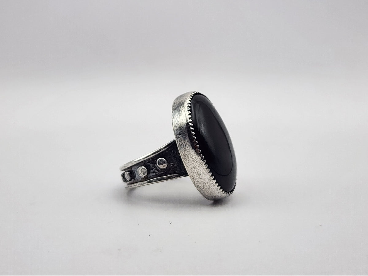 A side view of a sterling silver ring featuring a large, oval black onyx cabochon stone on a band with silver dots and heavy oxidation, photographed on a neutral light gray background.