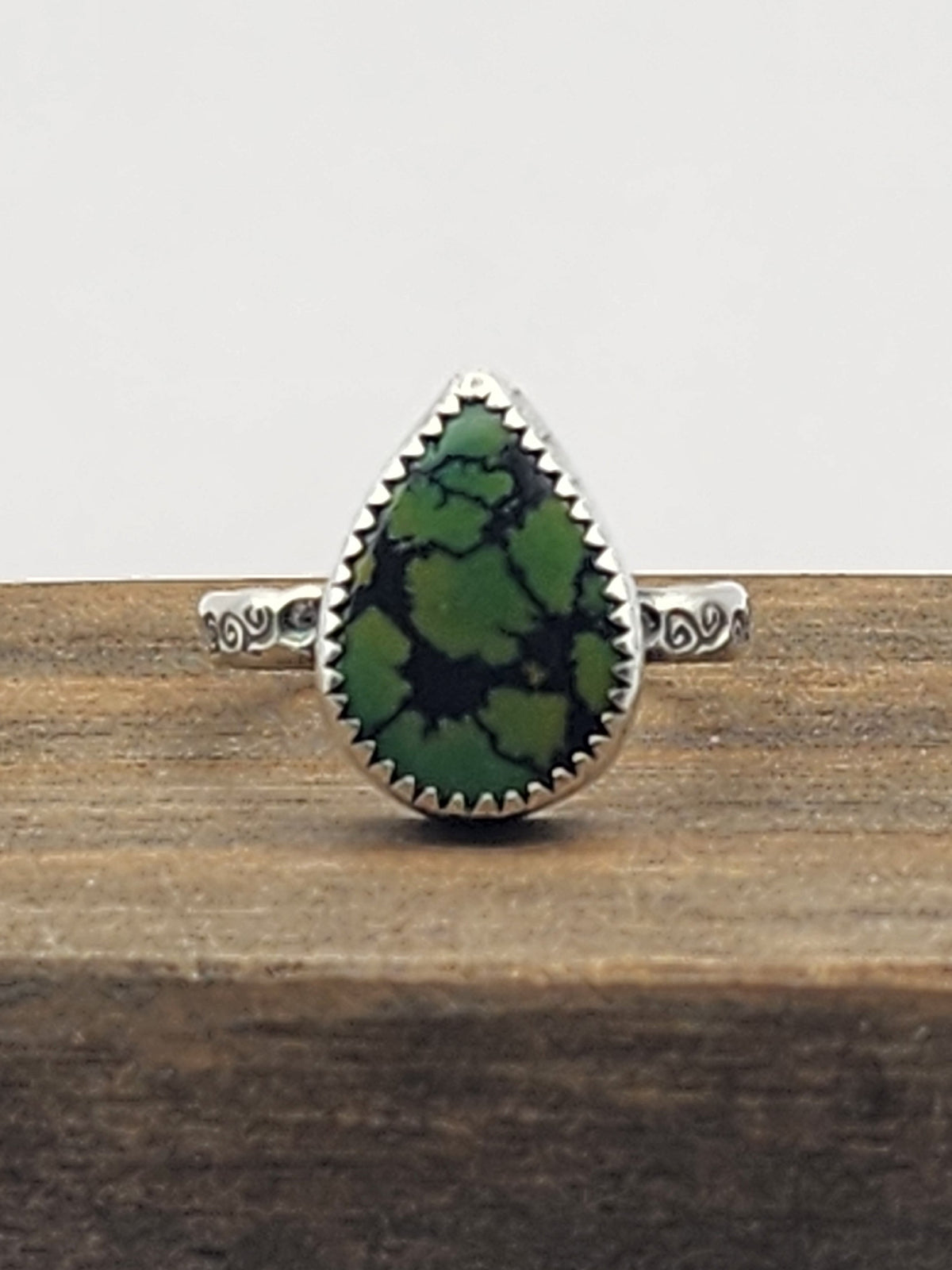 A sterling silver ring with swirl stamped band with a teardrop-shaped green turquoise gemstone with natural dark veining, displayed on a rustic wooden block against a light gray background.