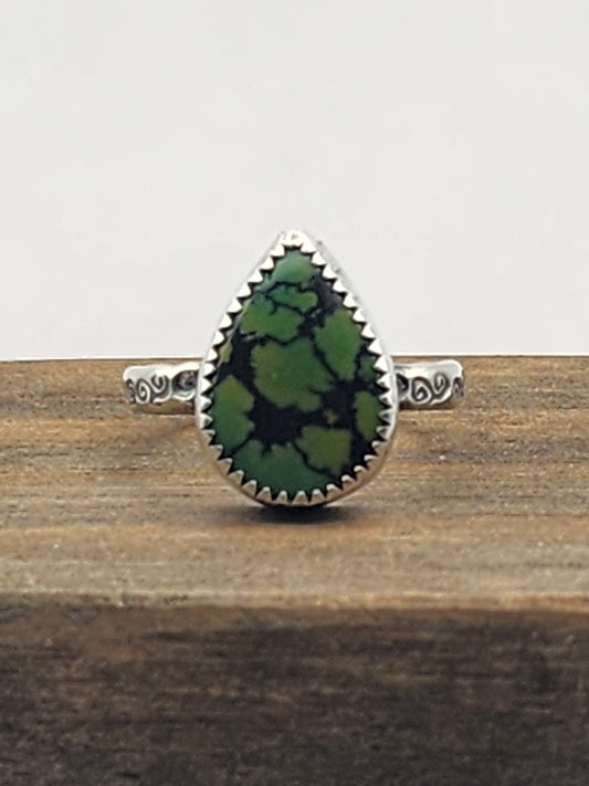 A sterling silver ring with swirl stamped band with a teardrop-shaped green turquoise gemstone with natural dark veining, displayed on a rustic wooden block against a light gray background.