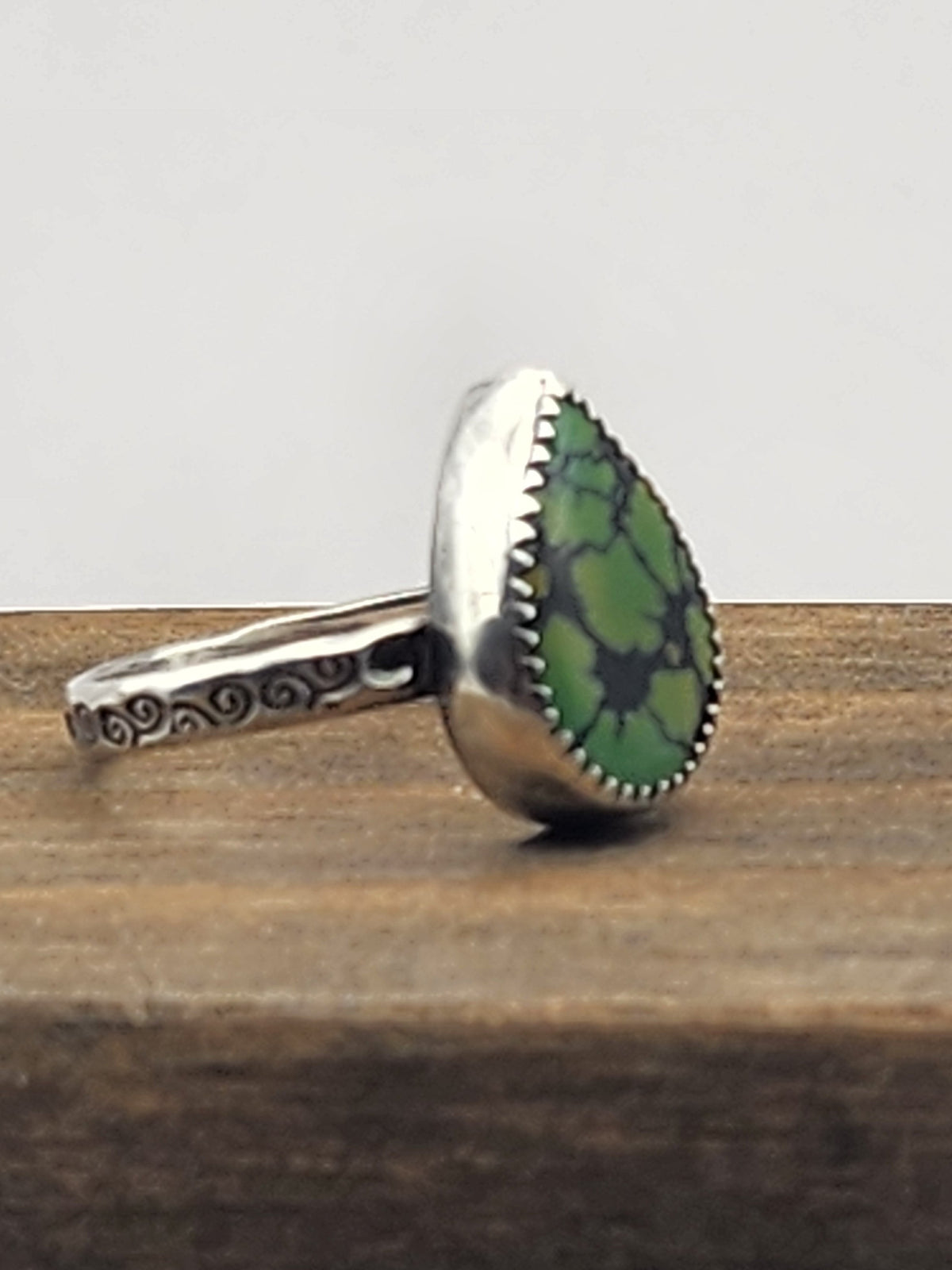 A side view of a sterling silver ring with swirl stamped band with a teardrop-shaped green turquoise gemstone with natural dark veining, displayed on a rustic wooden block against a light gray background.