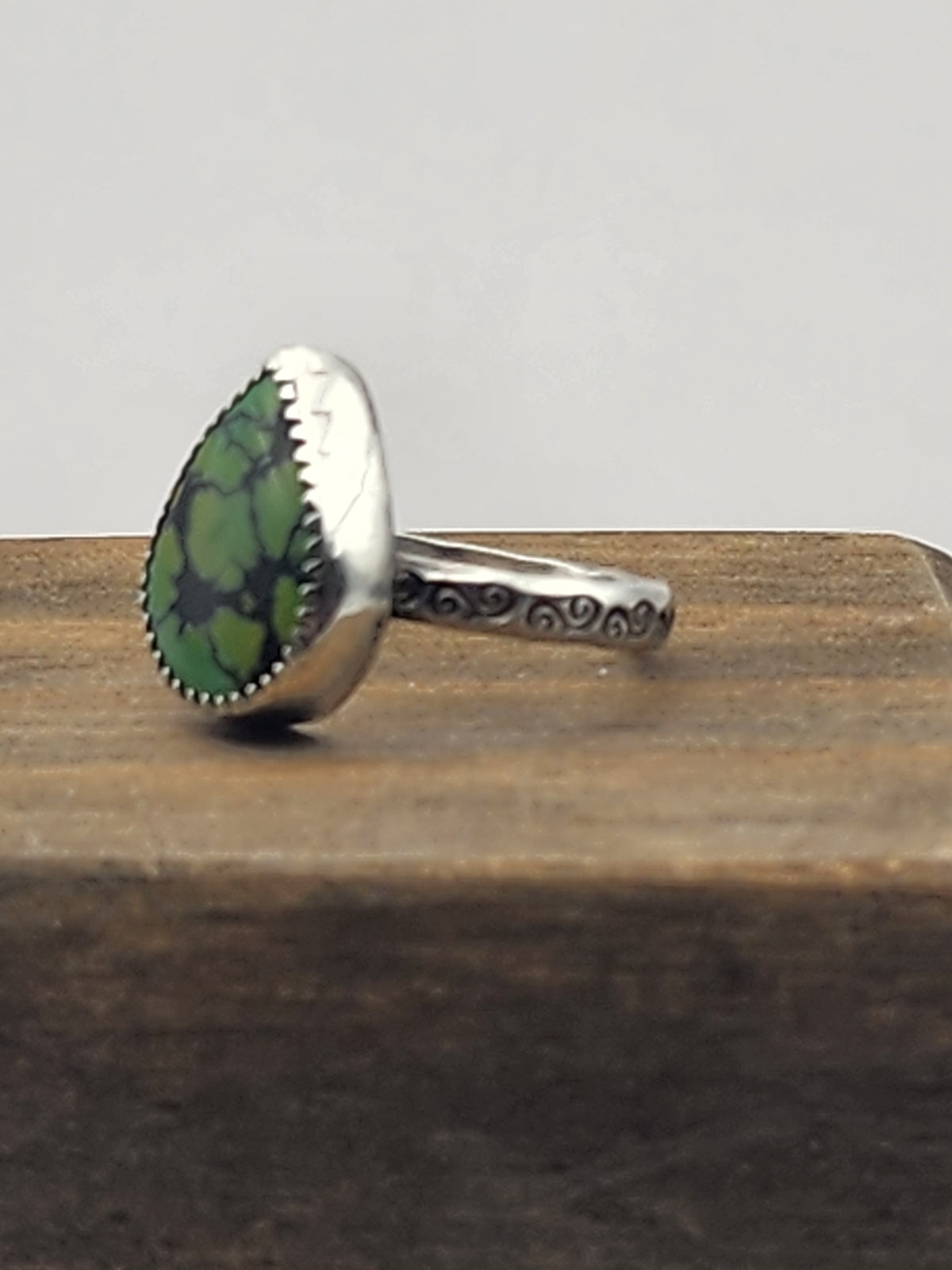 A side view of a sterling silver ring with swirl stamped band with a teardrop-shaped green turquoise gemstone with natural dark veining, displayed on a rustic wooden block against a light gray background.