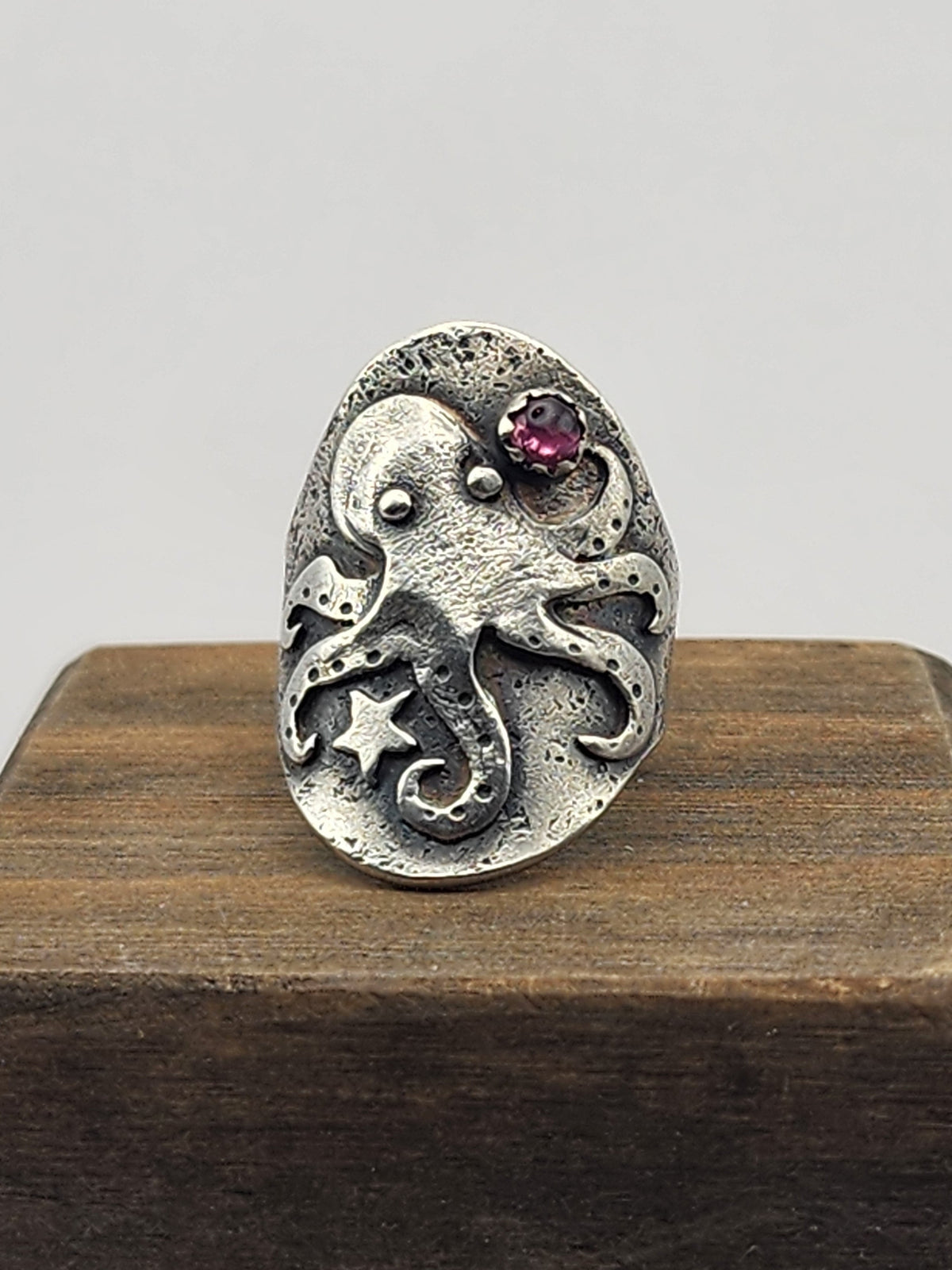 An oval sterling silver ring featuring a detailed octopus design with a small pink gemstone and star accents, displayed on a dark wooden block against a light gray background.