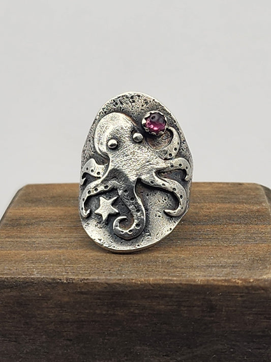 An oval sterling silver ring featuring a detailed octopus design with a small pink gemstone and star accents, displayed on a dark wooden block against a light gray background.