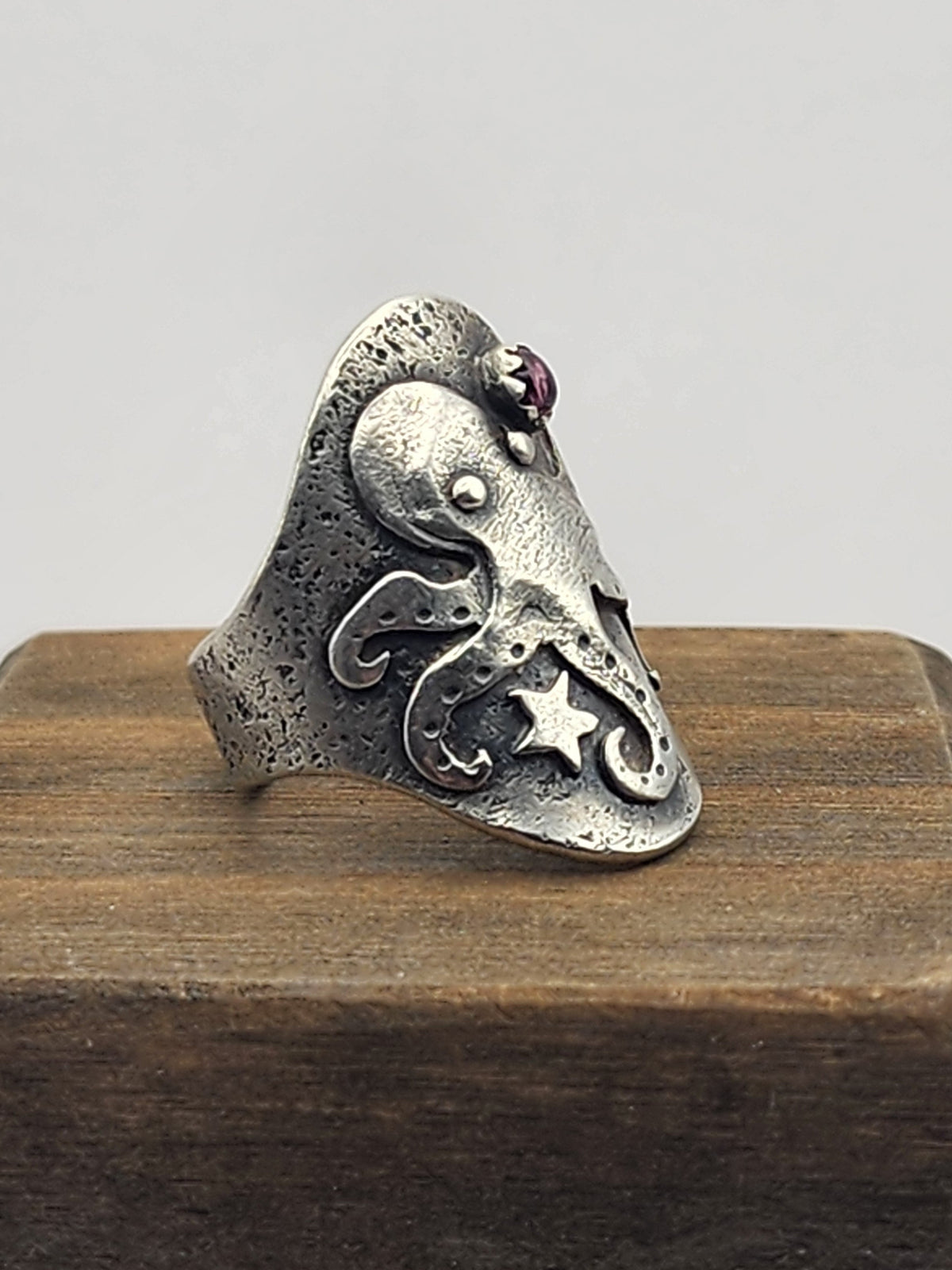 A sterling silver ring with an octopus, featuring a small pink gemstone beside its head, displayed on a wooden block against a light gray background.