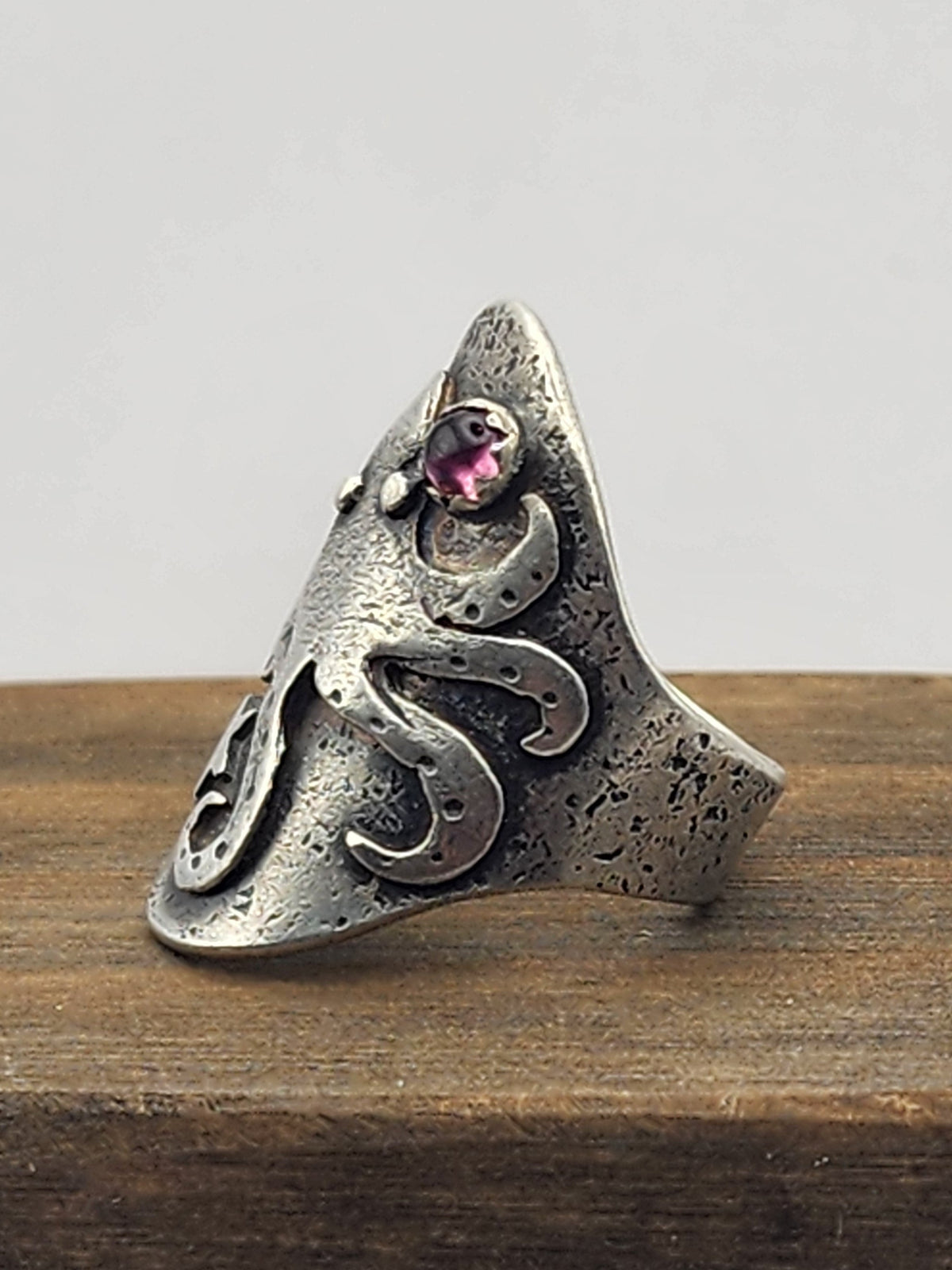 A side view of a textured sterling silver ring with an octopus , featuring a small pink gemstone, displayed on a wooden block against a light gray background.