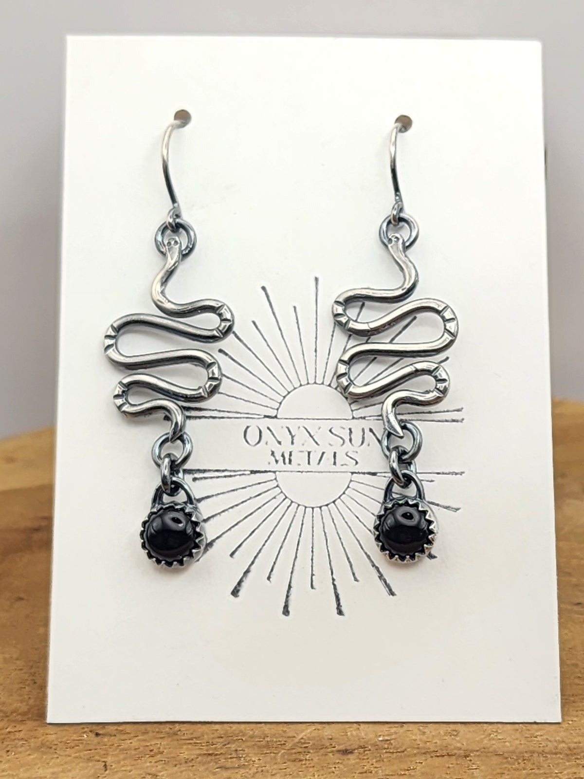 Pair of sterling silver dangle earrings with wire shaped into snakes and a small black onyx stone at the end of the snake. Earrings are on a white earring display card with a sun emblem and name stating onyx sun metal. card is sitting on wood display and a light grey background. 