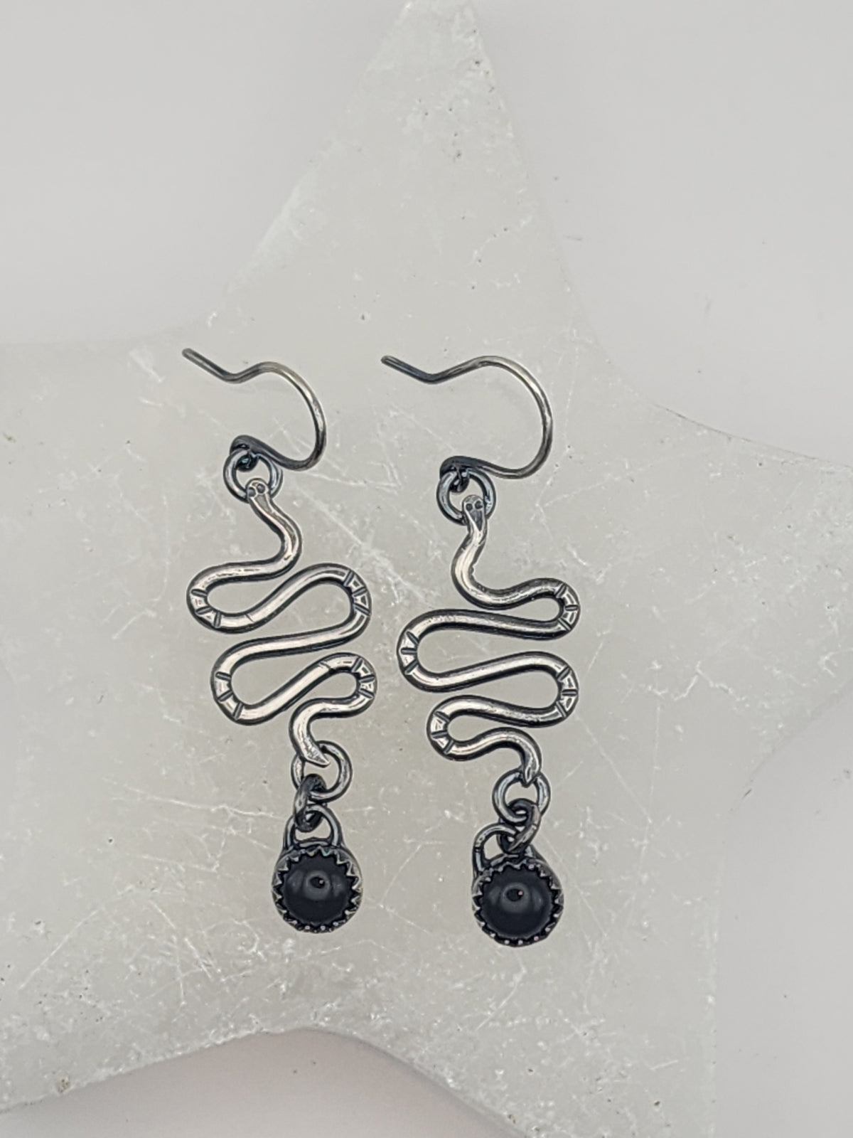 A pair of sterling silver dangle earrings with a snake-like design and dark round bead drops, displayed on a textured white star-shaped surface.