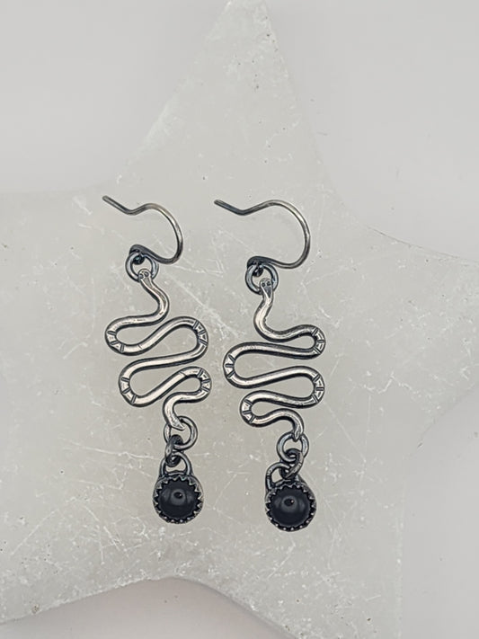 A pair of sterling silver dangle earrings with a snake-like design and dark round bead drops, displayed on a textured white star-shaped surface.