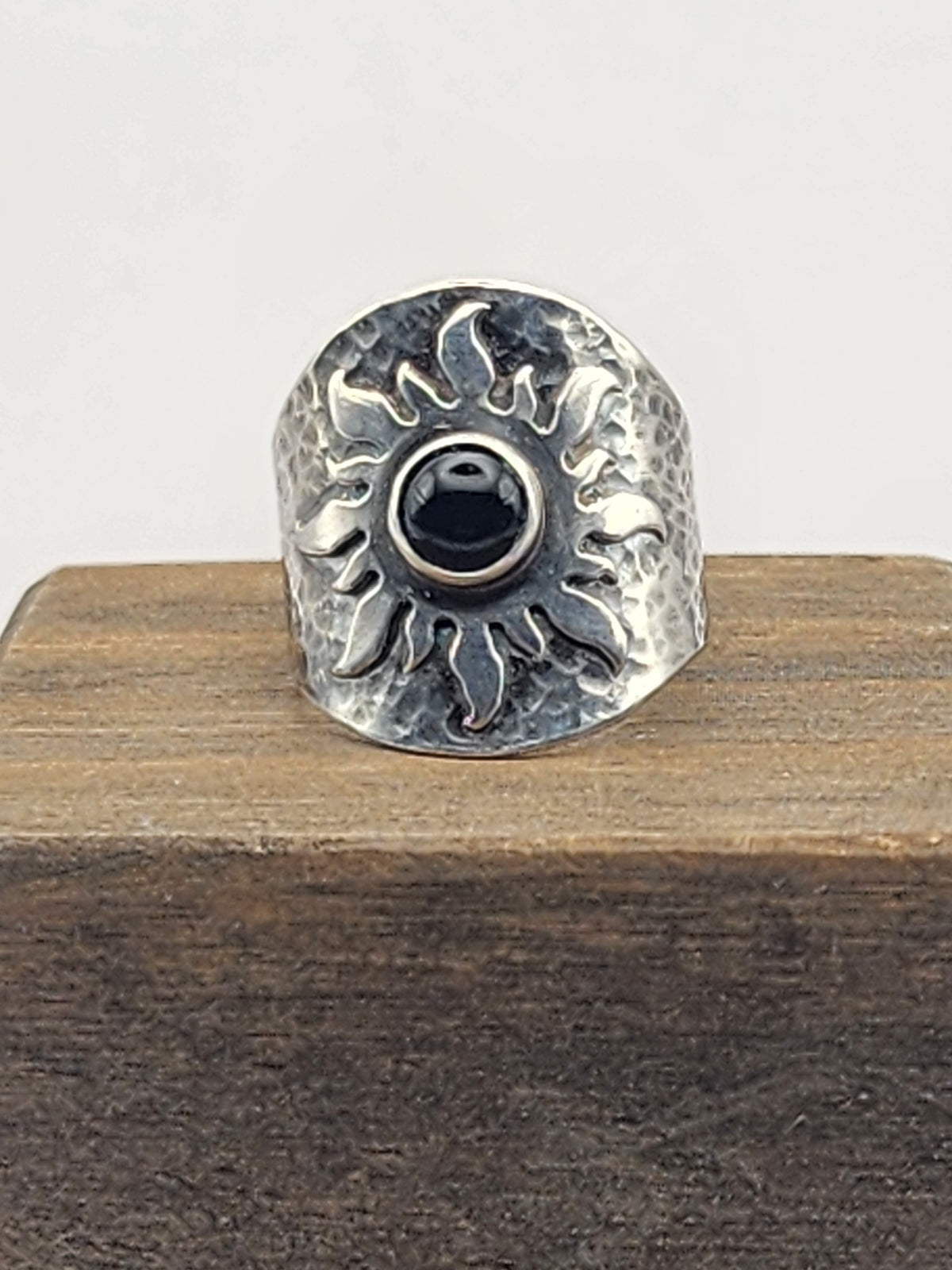 A hammered sterling silver ring with a central black round stone on a sun design, displayed on a wooden block against a light gray background.