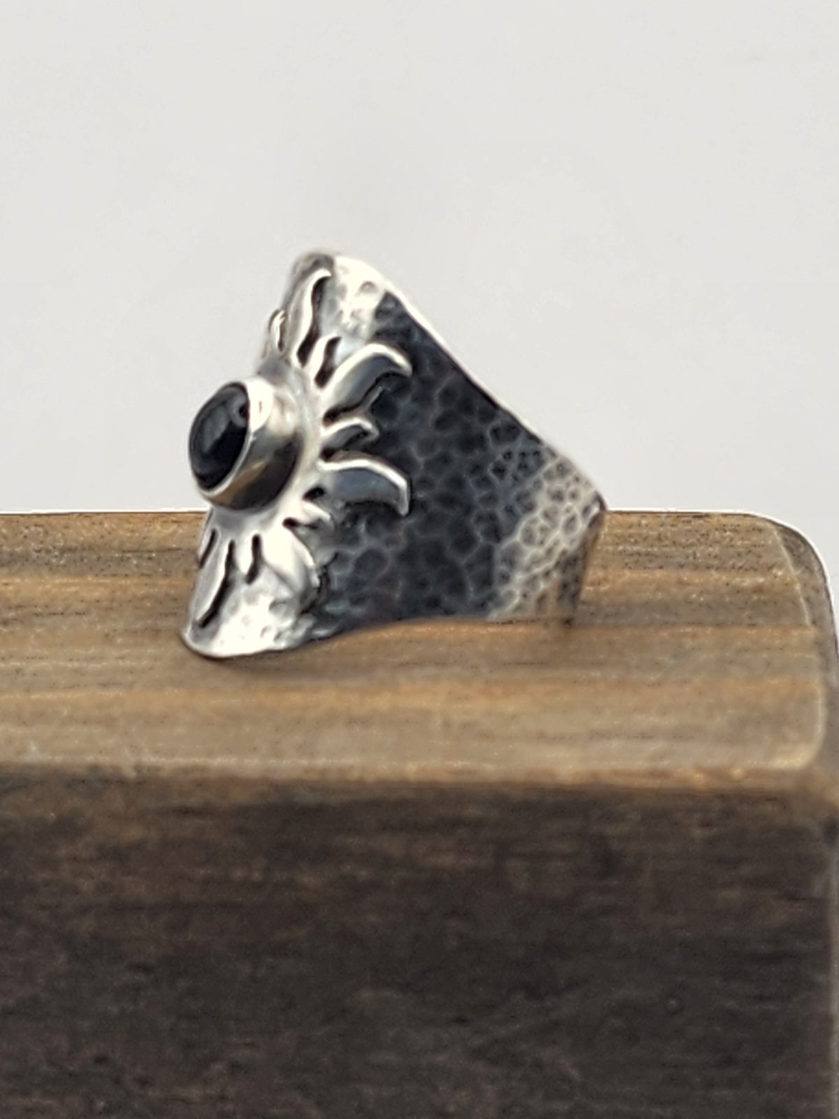 A side view of a hammered sterling silver ring with a central black round stone on a sun design, displayed on a wooden block against a light gray background.