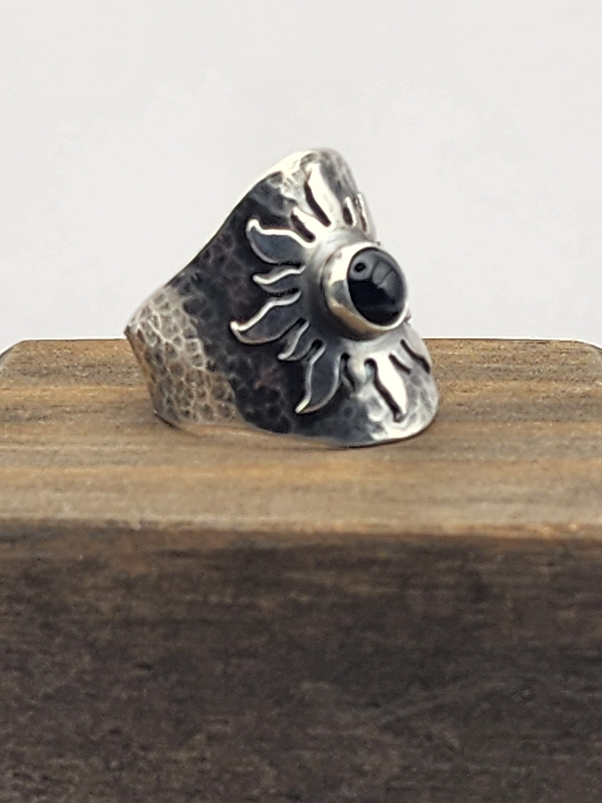 A side view of a hammered sterling silver ring with a central black round stone on a sun design, displayed on a wooden block against a light gray background.