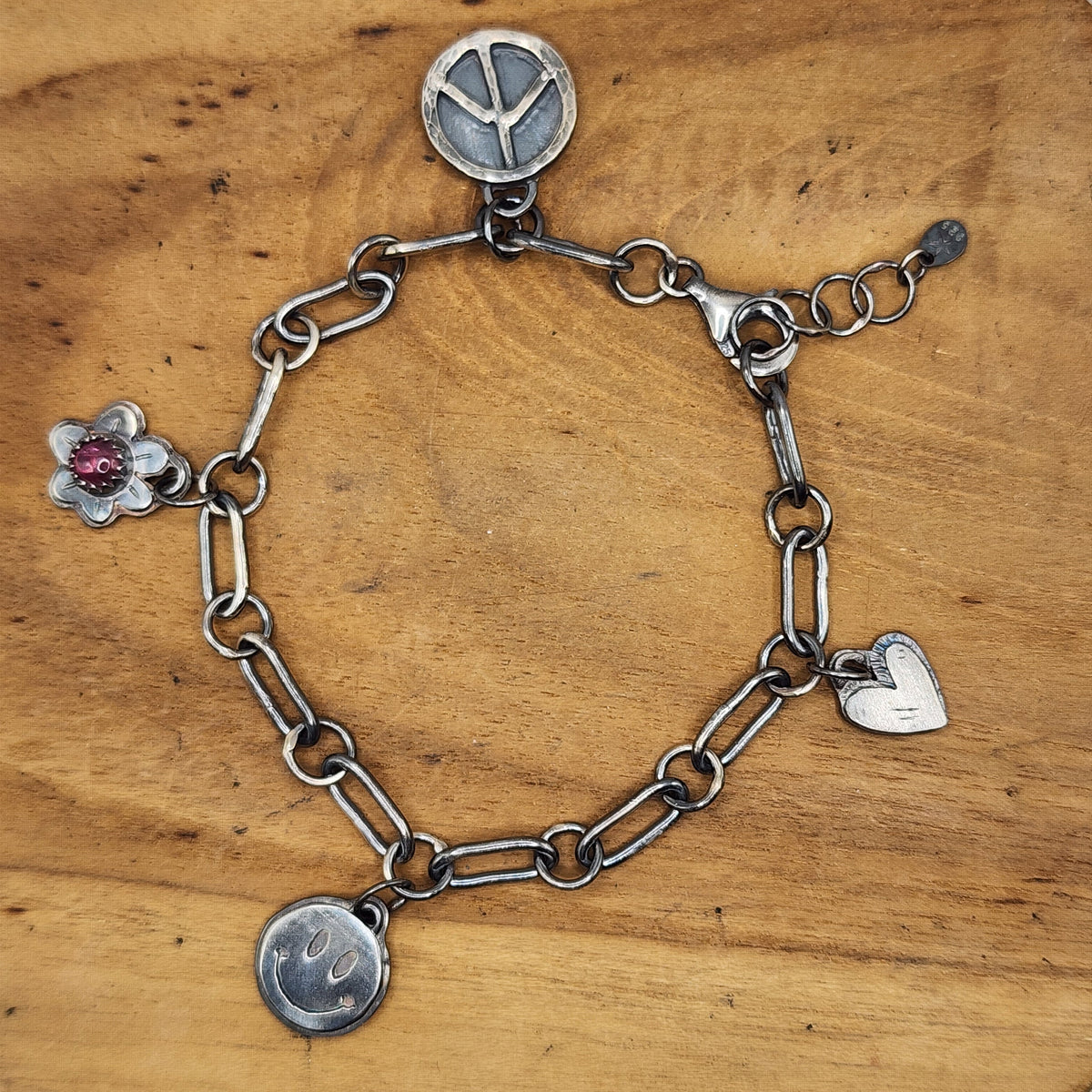 A  sterling silver chain bracelet with multiple charms, including a peace sign, a flower with a pink gemstone, a heart, and a smiley face, displayed on a wooden surface.