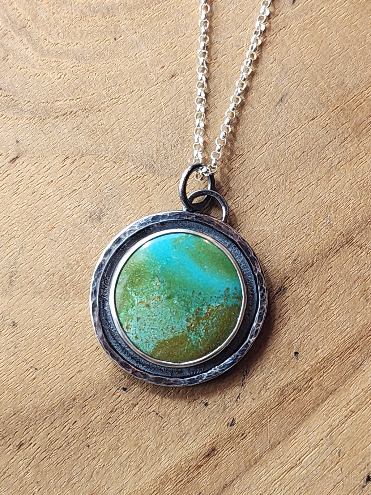Sterling silver pendant with round turquoise cabochon and textured border on a chain, sitting on a wooden background.