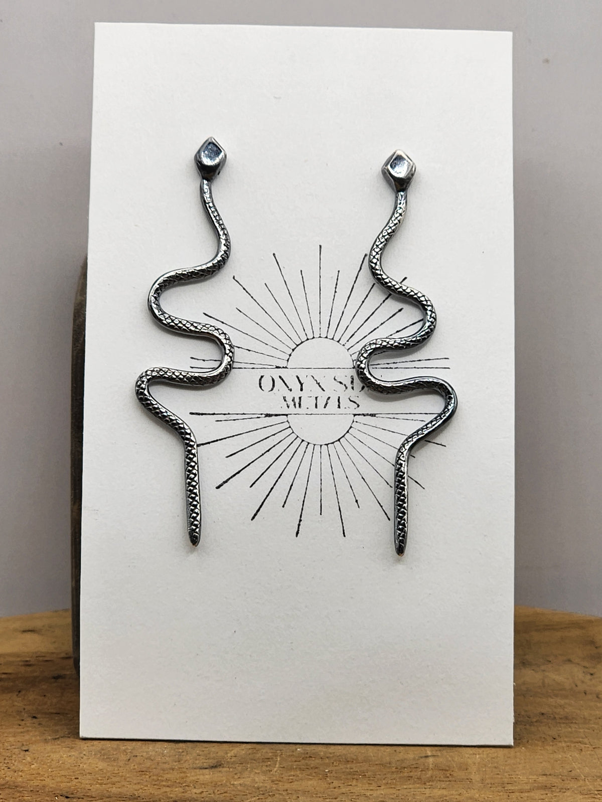 A pair of silver snake-shaped stud earrings, displayed on a white backing card featuring a black snake-and-sun illustration and the text 'ONYX METAL'.
