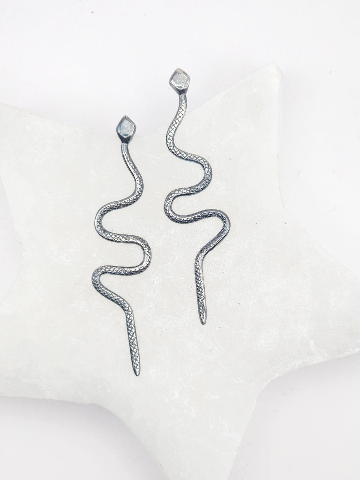 A pair of silver snake-shaped dangle earrings with textured scales, displayed on a white star-shaped jewelry stand against a clean white background.