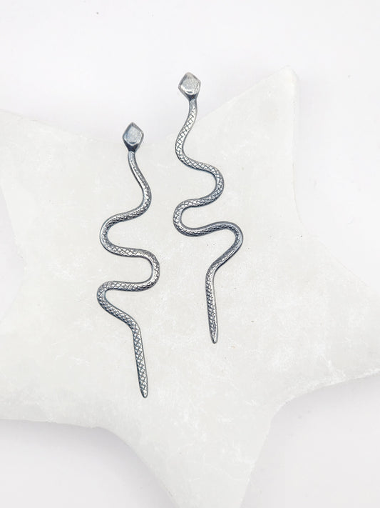 A pair of silver snake-shaped dangle earrings with textured scales, displayed on a white star-shaped jewelry stand against a clean white background.