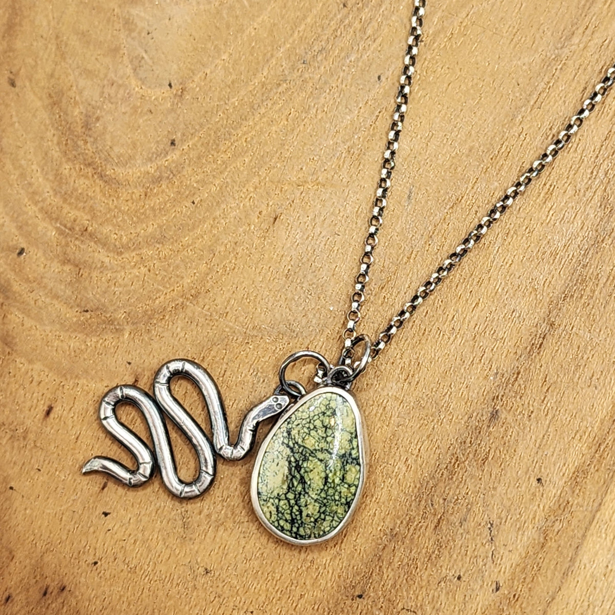 A sterling silver snake-shaped pendant necklace with a green marbled teardrop stone pendant, displayed on a light brown wooden surface.