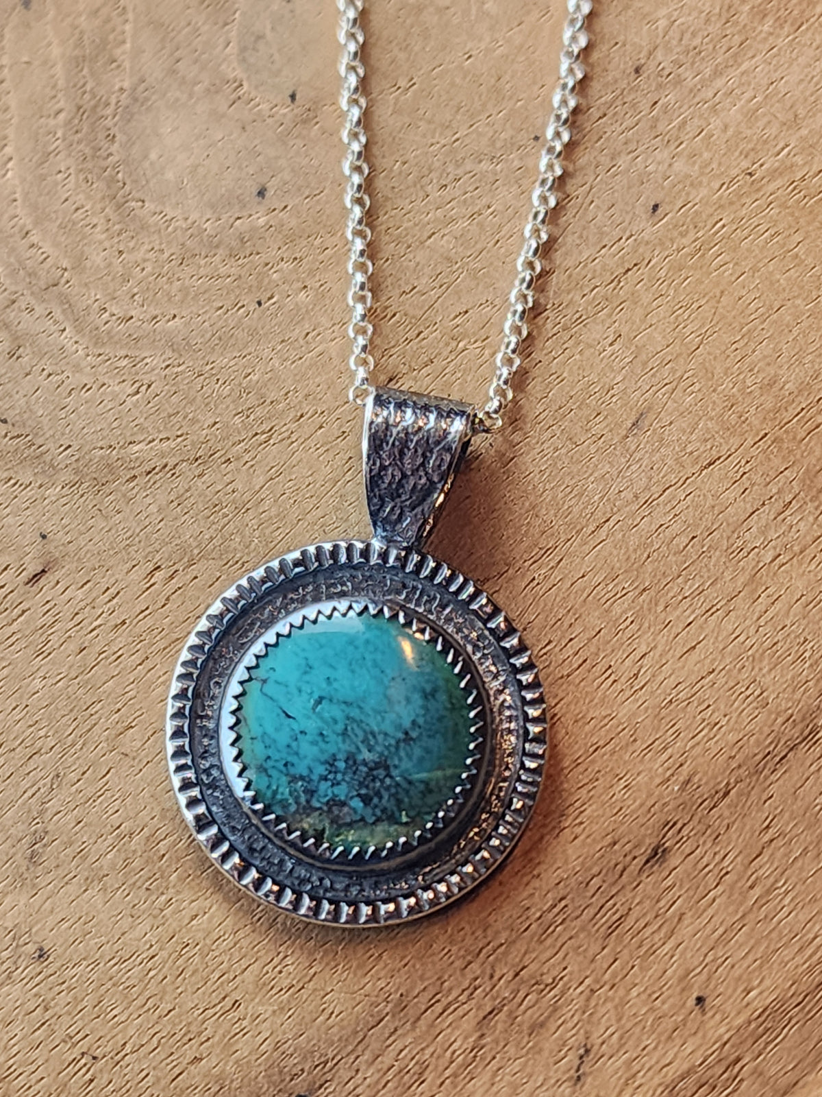Sterling silver pendant with round turquoise cabochon and textured border, on a silver chain, sitting on a wooden platform.