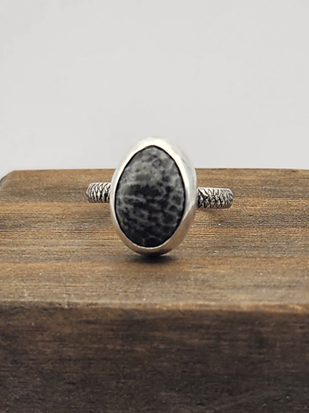 A sterling silver ring with a textured band, dark gray and black speckled oval stone, displayed on a dark wooden block against a light gray background.