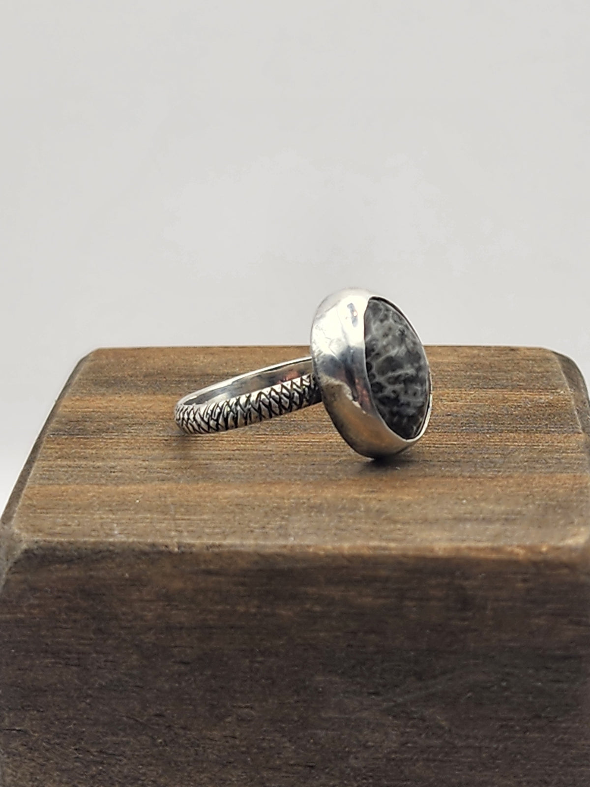 A side view of a sterling silver ring with a textured band, dark gray and black speckled oval stone, displayed on a dark wooden block against a light gray background.