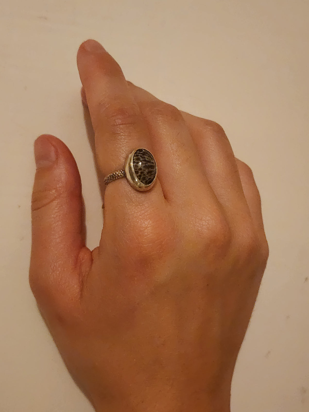 Sterling silver ring with textured band and specked grey and black stone on pointer finger of a hand on an off white background.