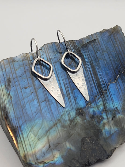 A pair of silver geometric dangle earrings with open diamond shape and pointed triangular drops, displayed on a polished blue labradorite stone against a light gray background.
