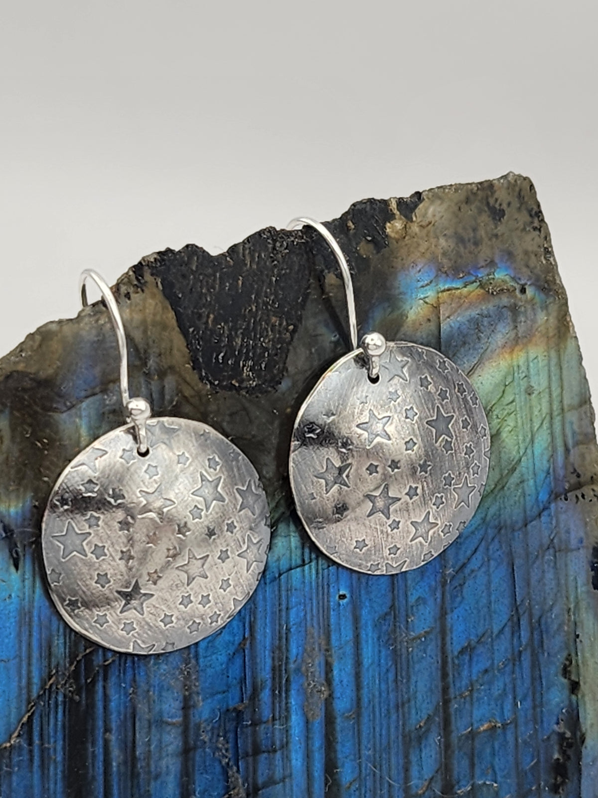 A pair of sterling silver silver disc earrings imprinted with stars, hanging from silver fishhook wires, displayed on a labradorite stone against a neutral gray background.