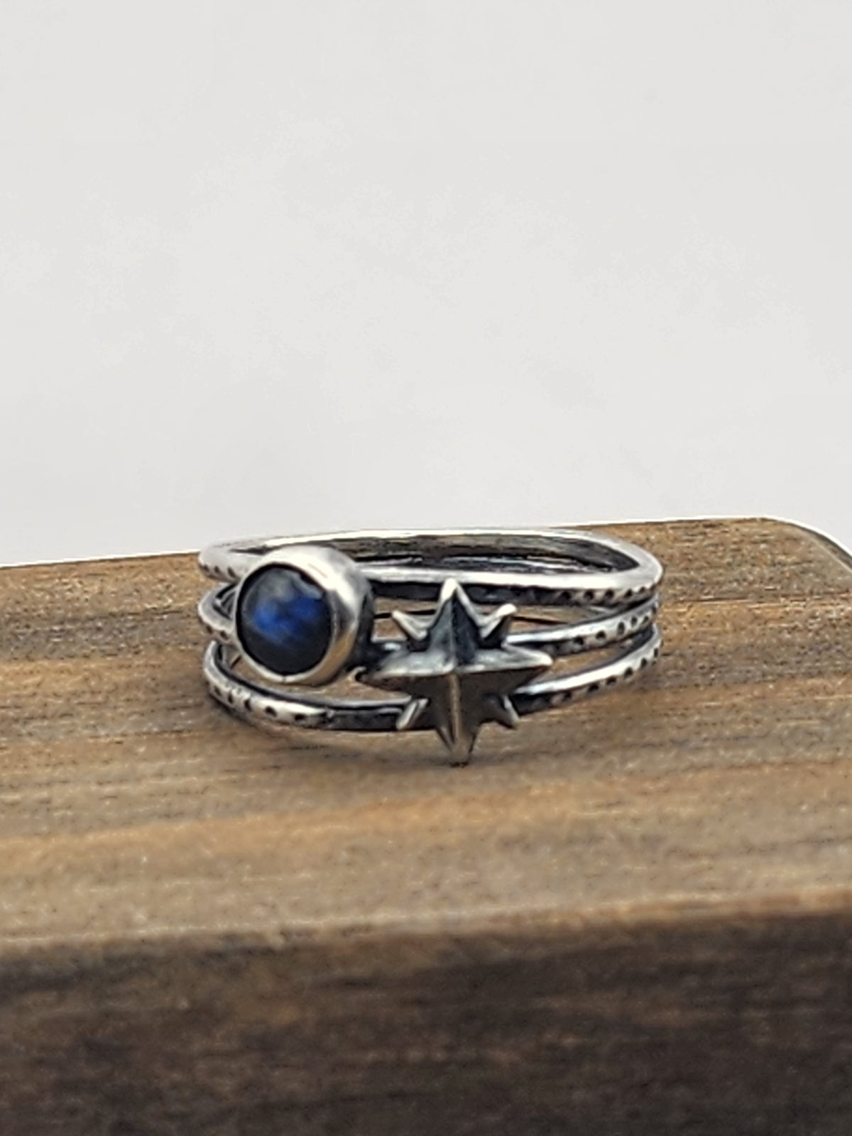 A sterling silver three-band textured ring featuring a starburst design and a round labradorite cabochon stone, displayed on a wooden block against a neutral light gray background.