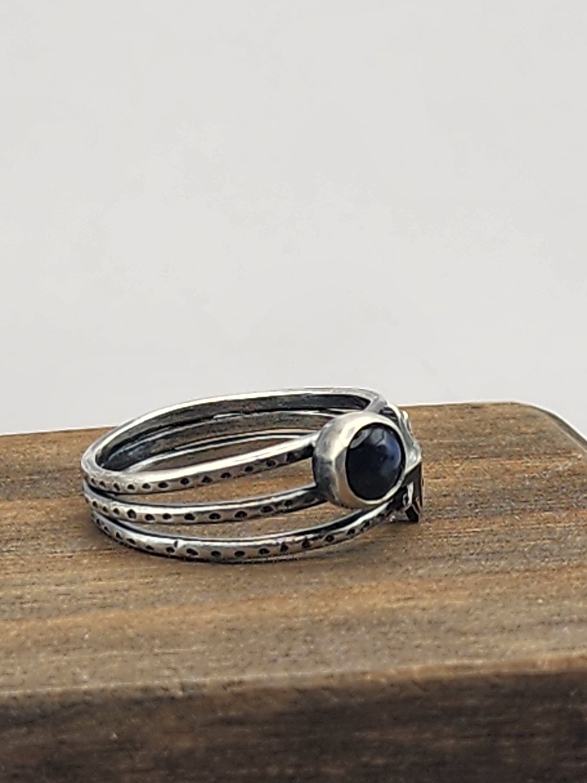 A side view of a sterling silver three-band textured ring featuring a starburst design and a round labradorite cabochon stone, displayed on a wooden block against a neutral light gray background.