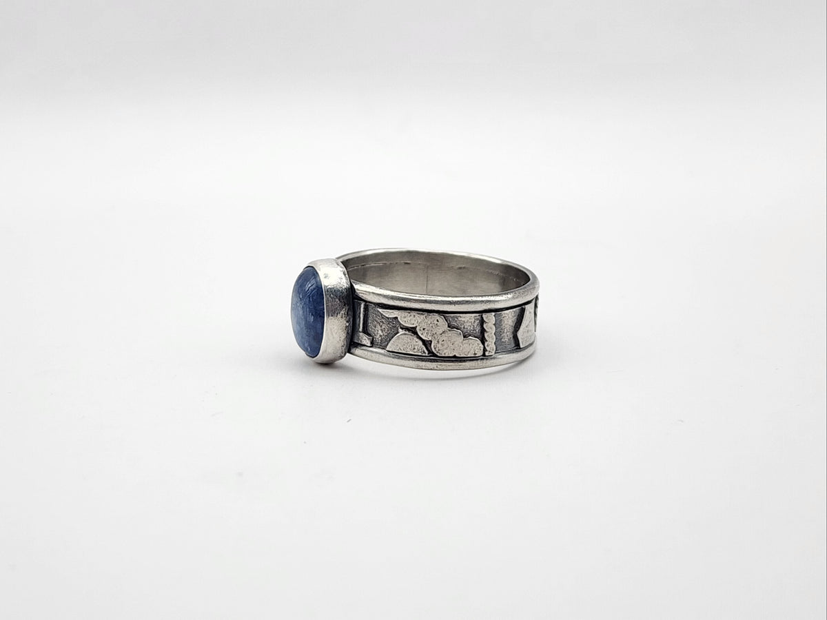 A side view of a sterling silver ring featuring a central oval blue stone set on a heavily textured and oxidized band, set against a plain light gray background.