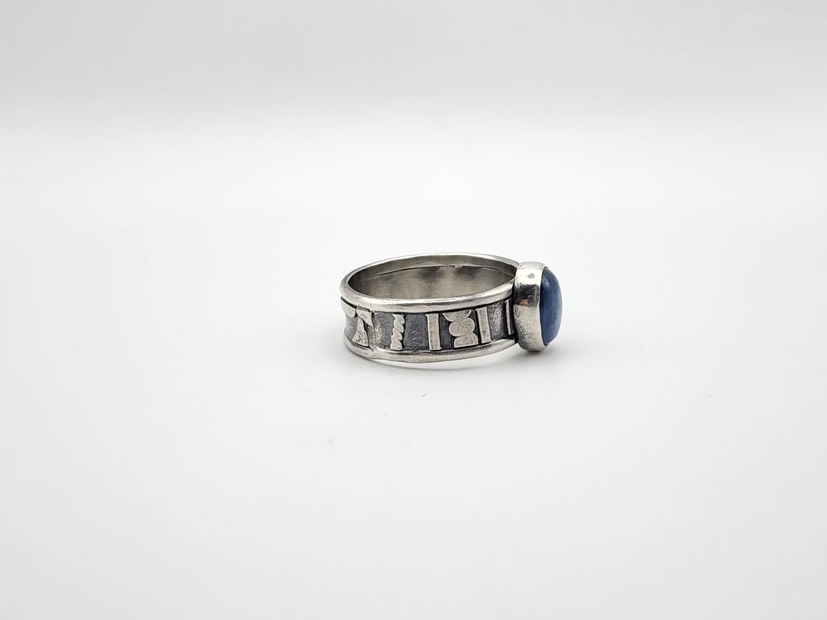 A side view of a sterling silver ring featuring a central oval blue stone set on a heavily textured and oxidized band, set against a plain light gray background.