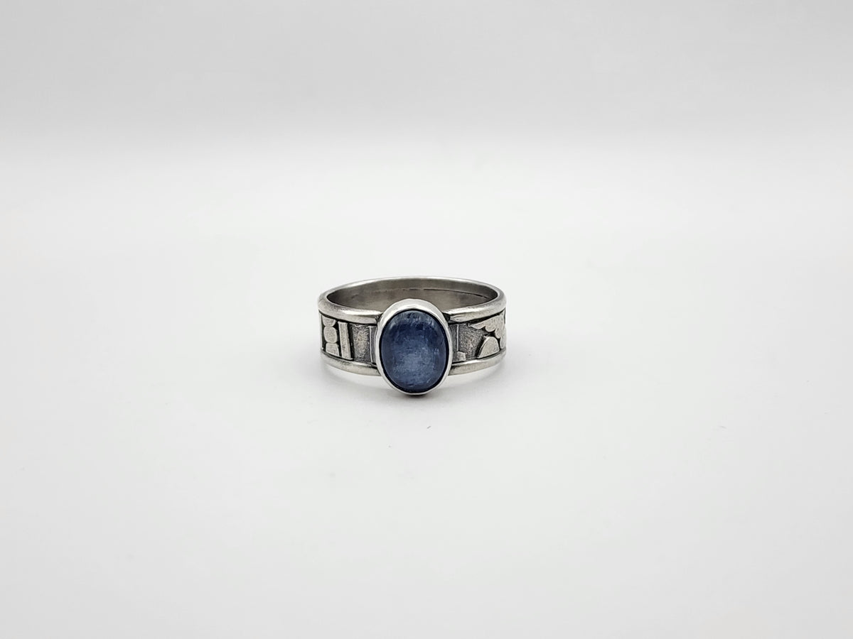 A sterling silver ring featuring a central oval blue stone set on a heavily textured and oxidized band, set against a plain light gray background.