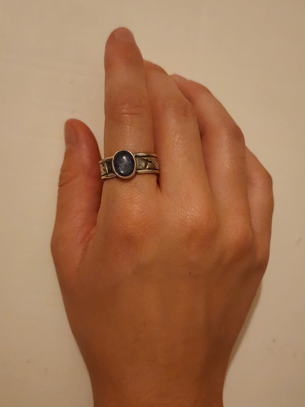 A sterling silver  ring with an oval dark blue cabochon stone and heavily textured wide band, worn on a person's pointer finger against a plain beige background.