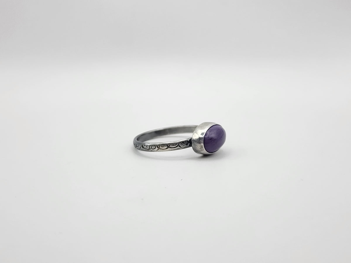 A side view of a sterling silver ring with an oval purple cabochon gemstone on a stamped ring band, photographed on a plain light gray background.