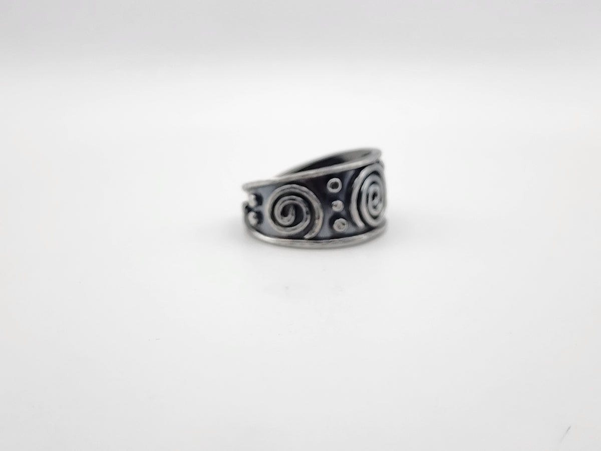 A sterling silver ring with a wide band featuring spiral and dot patterns, shown on a plain light gray background.