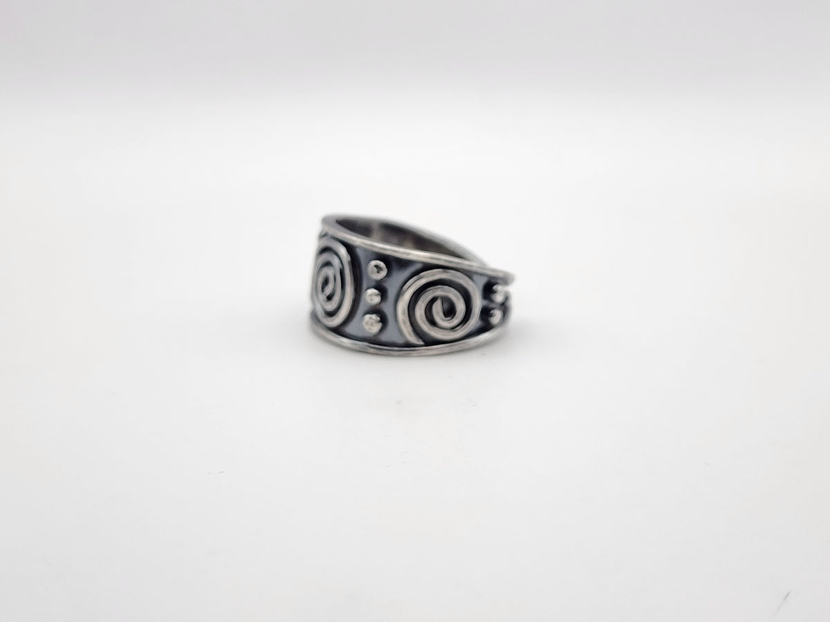 A sterling silver ring with a wide band featuring raised spiral and small circular dot details, set against a plain white background.