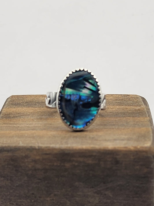A sterling silver ring with an oval abalone cabochon, displayed on a dark wooden block against a light gray background.