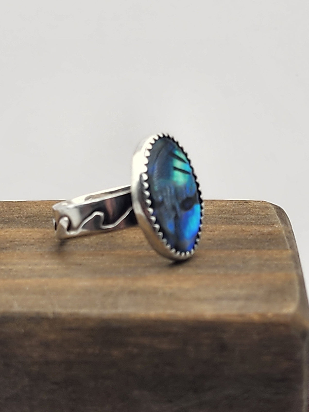 A sterling silver ring with a wave design and an oval blue abalone shell cabochon, displayed on a dark wood block against a light gray background.