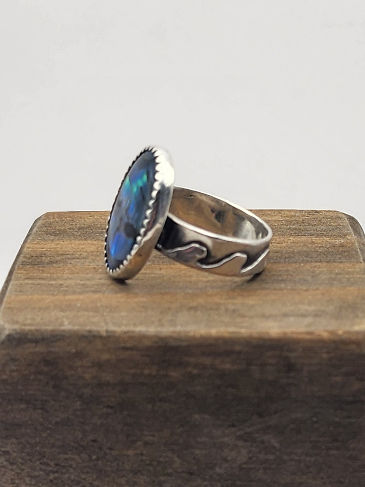 A sterling silver ring with a wave design and an oval blue abalone shell cabochon, displayed on a dark wood block against a light gray background.