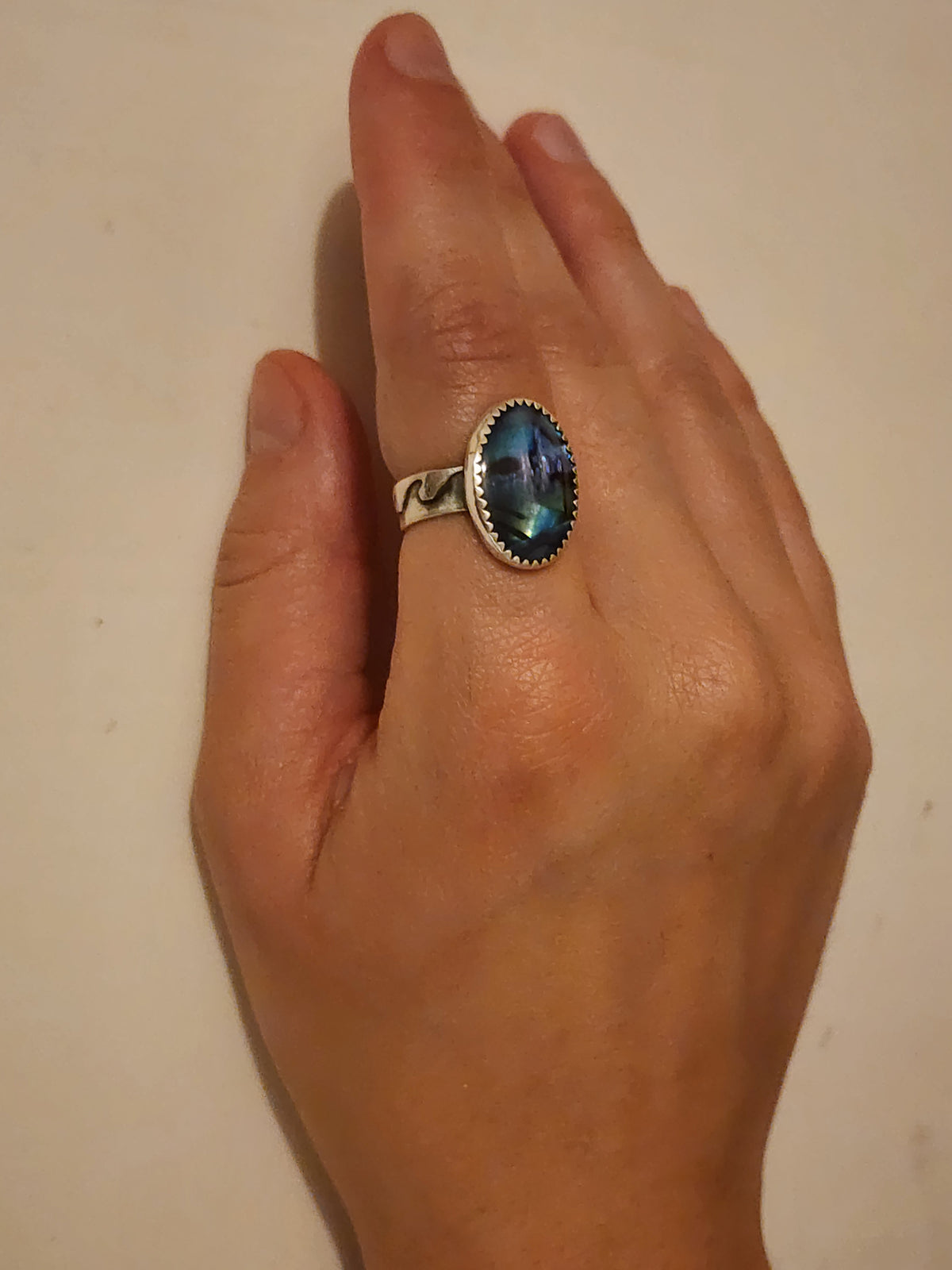 A hand wearing a sterling silver ring with an oval blue abalone cabochon, set against a plain off-white background.