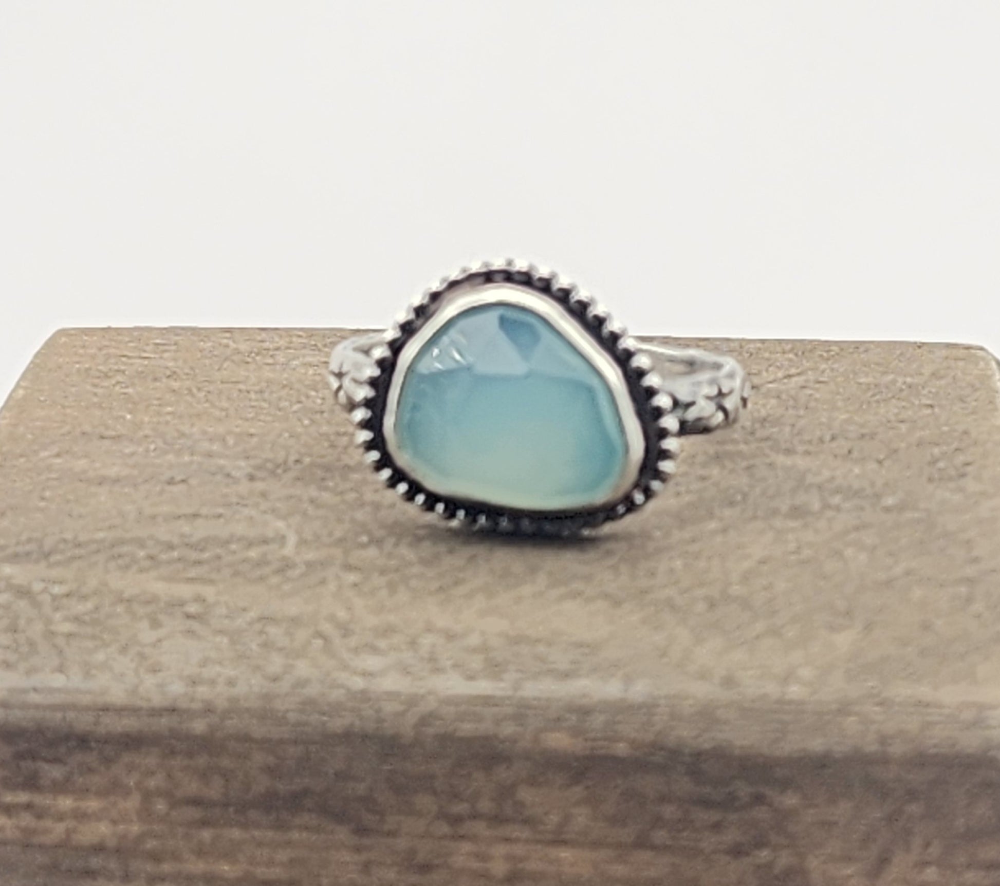 A silver ring with a large, irregular light blue stone, displayed on a textured brown wooden block against a white background.