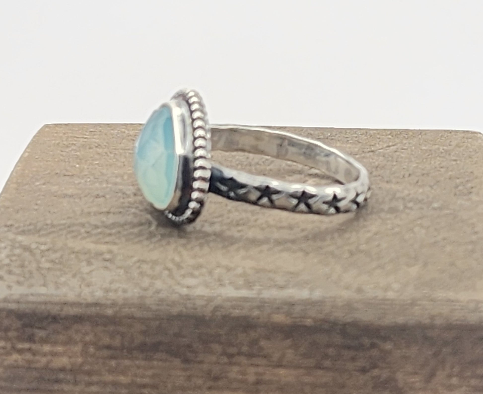 Side view of a sterling silver ring with a round, light blue cabochon gemstone and a band featuring star-stamped details, displayed on a textured wooden block.