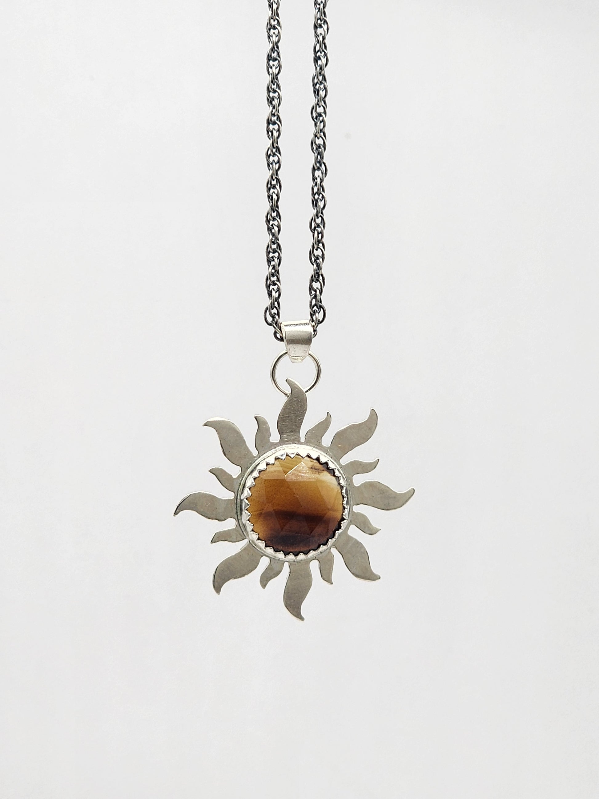 A silver-toned sun pendant necklace featuring a central oval brown and amber montana agate stone, suspended from a delicate silver chain against a plain white background.